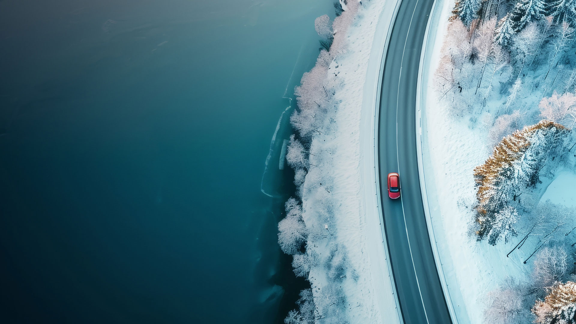 In a snowy landscape, a red car winds its way along a road bordering a body of water.