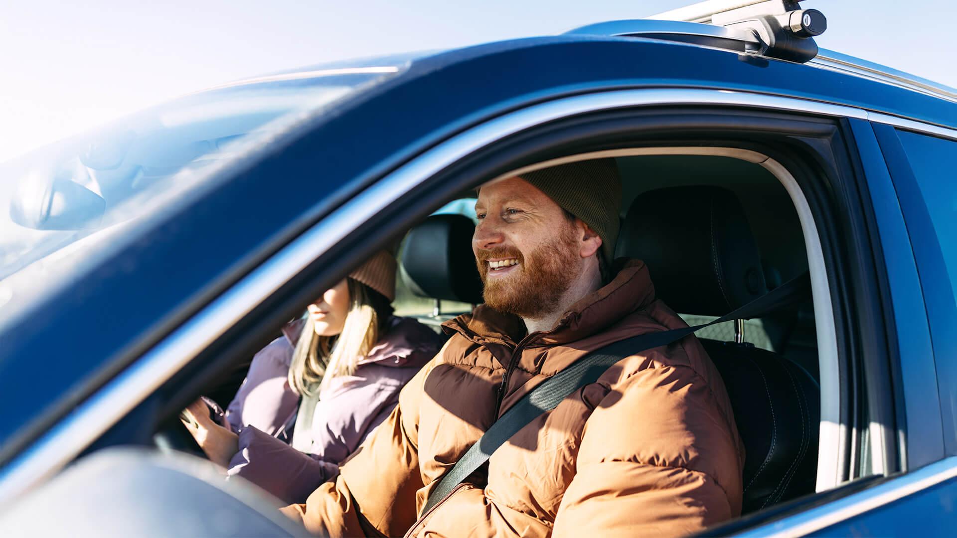 Un homme et une femme sourient, à bord de leur véhicule. A man and a woman sit smiling in their vehicle.